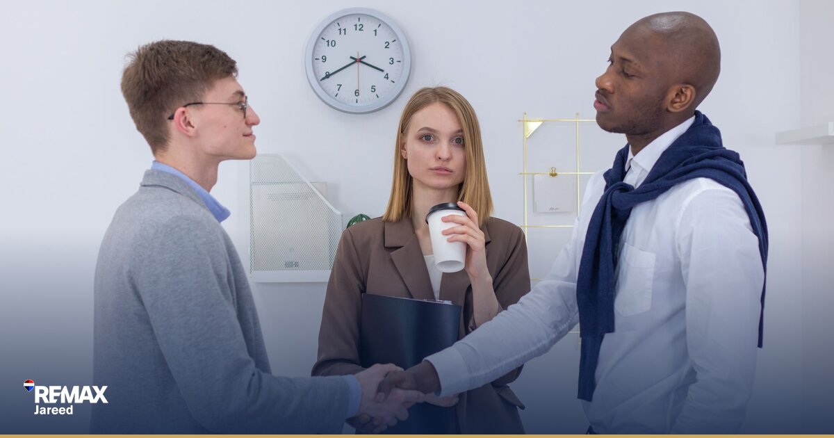 Two professionals shaking hands across a desk in a modern office, symbolizing a career partnership and commission agreement in real estate